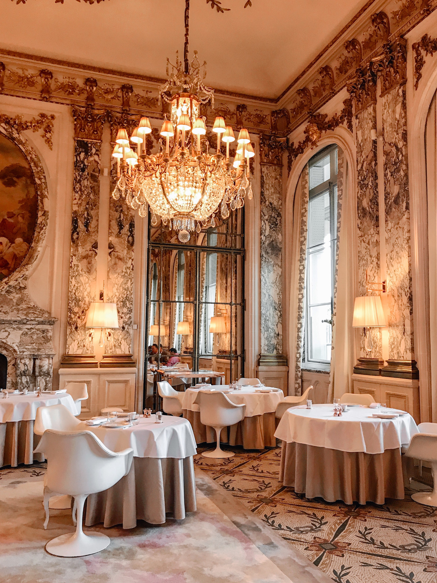Restaurant room with a focus on a large chandelier lighting the gold ornaments on the walls. At Le Meurice, Paris.