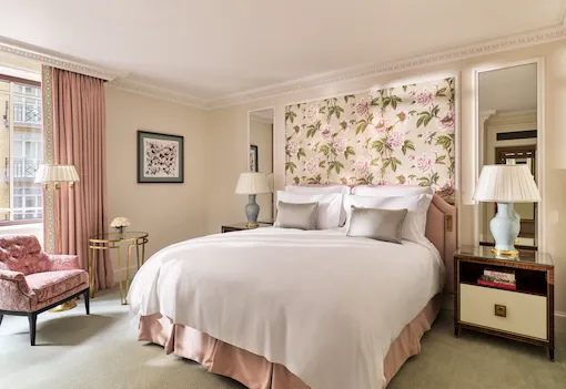 The Superior Room at The Dorchester, showcasing a king-sized bed framed by botanical floral wall coverings, blush-pink upholstery, classic bedside tables and soft natural light from the window.