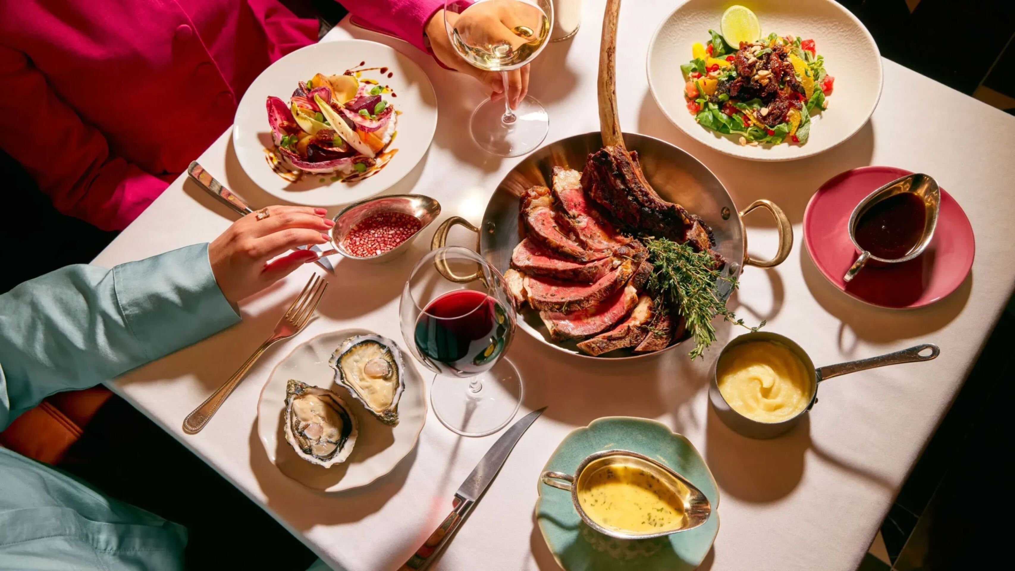 Dining table at The Grill at The Dorchester, showcasing a sliced Tomahawk steak served on the bone, accompanied by oysters, seasonal salads, rich sauces and glasses of red wine in an elegant sharing-style feast.