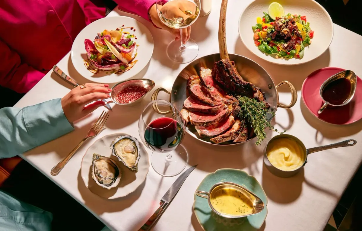 Dining table at The Grill at The Dorchester, showcasing a sliced Tomahawk steak served on the bone, accompanied by oysters, seasonal salads, rich sauces and glasses of red wine in an elegant sharing-style feast.