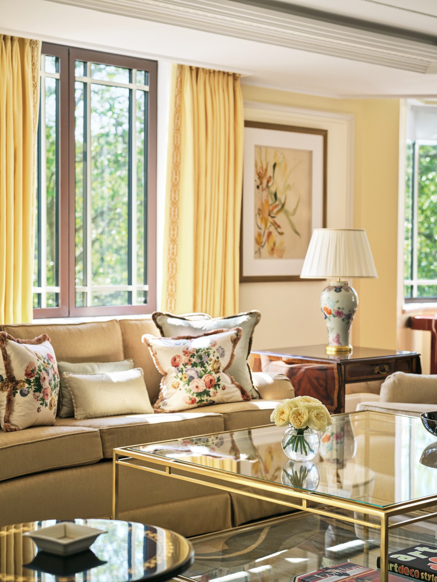 The Dorchester Suite living room at The Dorchester, showcasing comfortable lounge seating, floral accent cushions, a glass coffee table, soft yellow drapery and large windows framing views of Hyde Park and Park Lane.