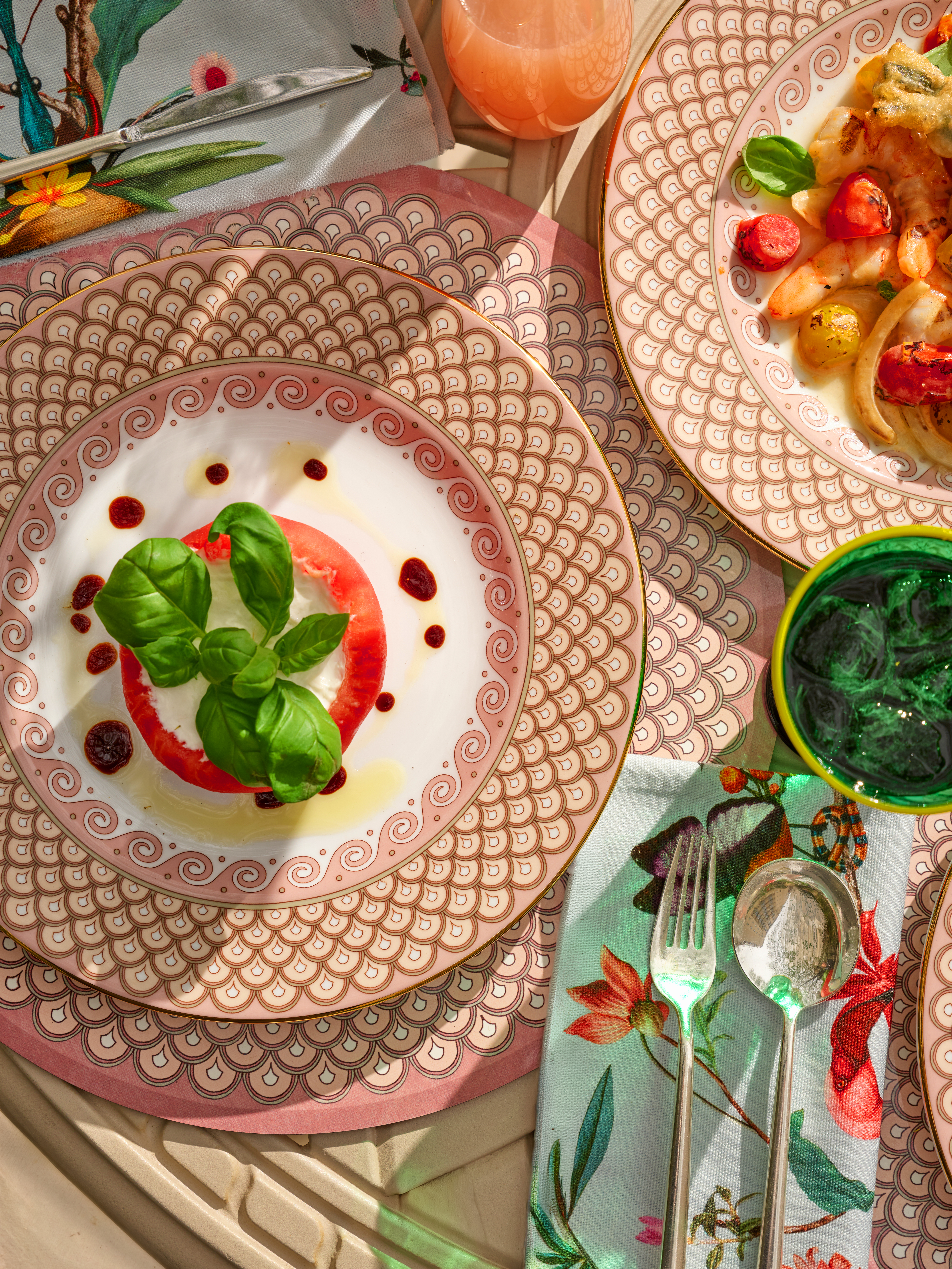 La DoubleJ Summer Garden tablescape featuring layered decorative plates, fresh seasonal dishes, green glassware, and floral accents in a vibrant celebration of outdoor dining.