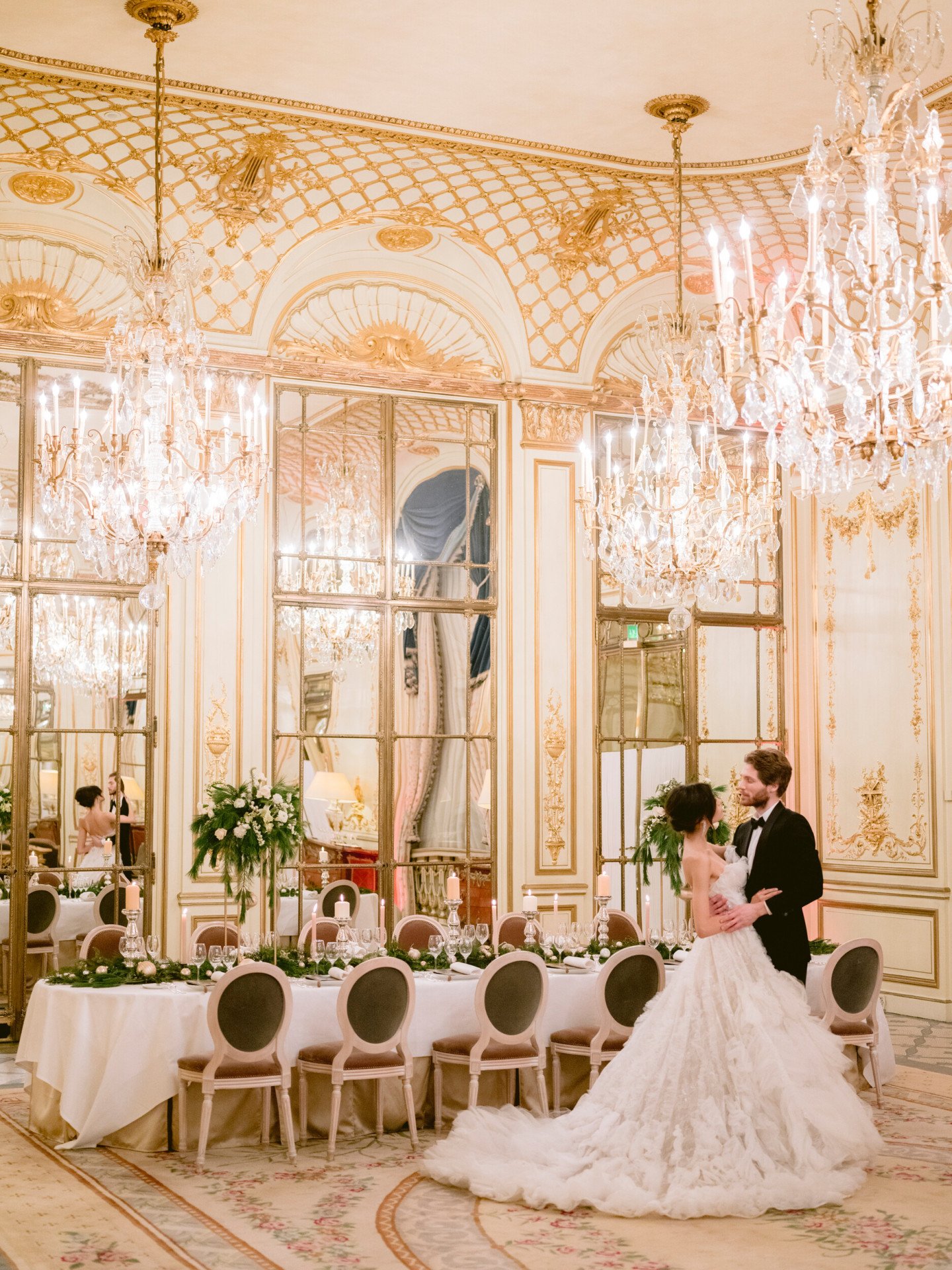 Wedding couple standing in front of a festive set table in Salon Pompadour. Green and white flower arrangements are on the table. The bride is looking at her husband, at Le Meurice, Paris