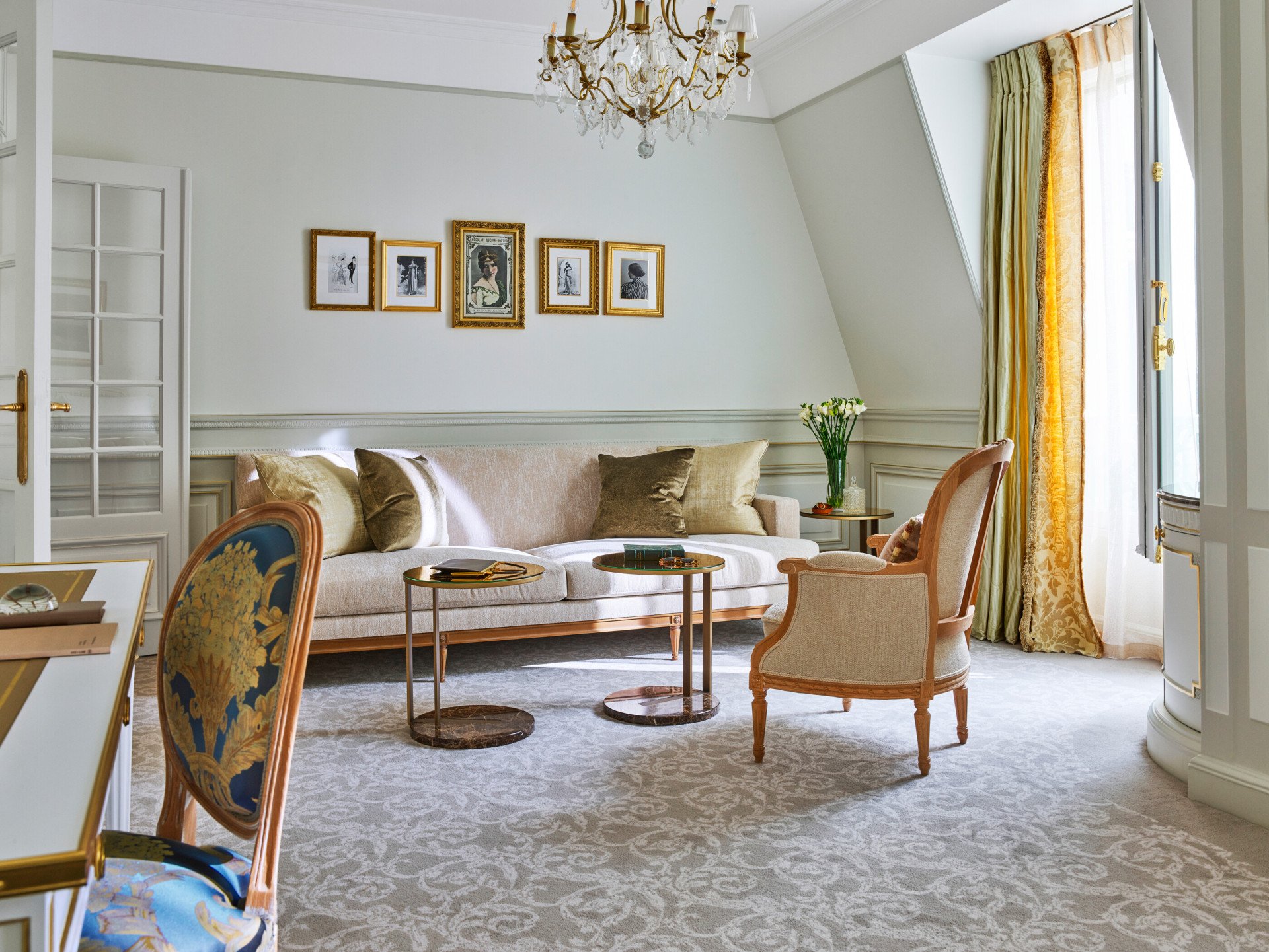 Élégant salon avec un canapé beige, des coussins décoratifs et deux tables rondes. Un lustre doré est suspendu au plafond. Des œuvres d'art encadrées ornent les murs.