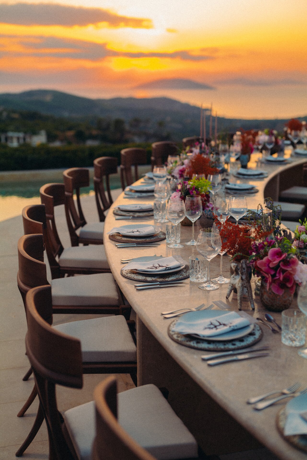 Elegante jantar ao ar livre com uma longa mesa, decorada com flores coloridas e louça requintada, tendo como pano de fundo um pôr do sol vibrante sobre colinas ondulantes.