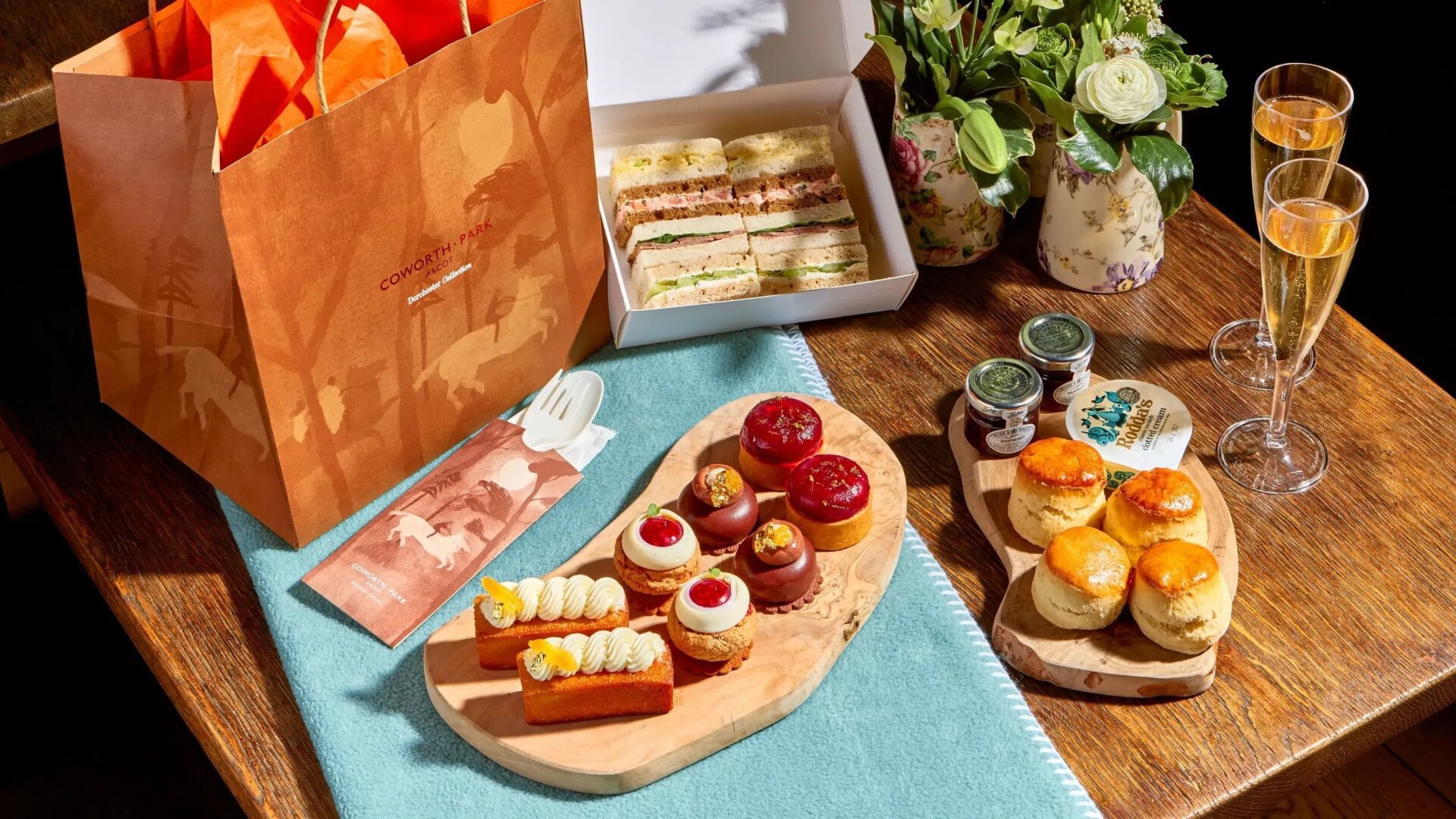 Takeaway meadow afternoon tea with finger sandwiches in box, summer pastries, scones, cream and jam and champagne layed out on picnic blanket with takeaway bag for the Royal Windsor Flower Show