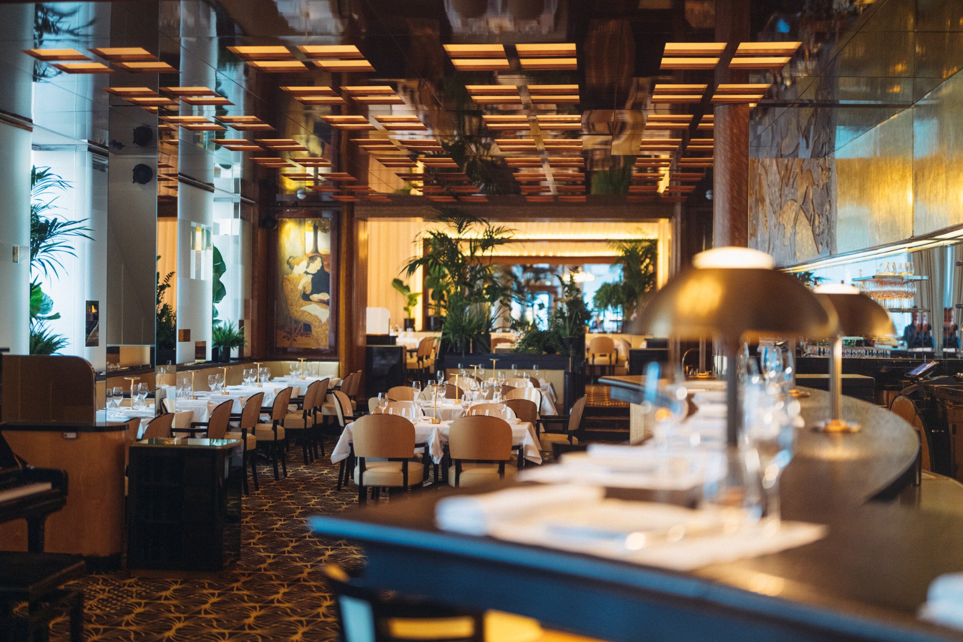 Intérieur élégant avec une ambiance chaleureuse et accueillante. Les tables sont dressées avec des nappes blanches impeccables, entourées de chaises beiges. Des plantes vertes et un éclairage tamisé renforcent l'atmosphère cosy.