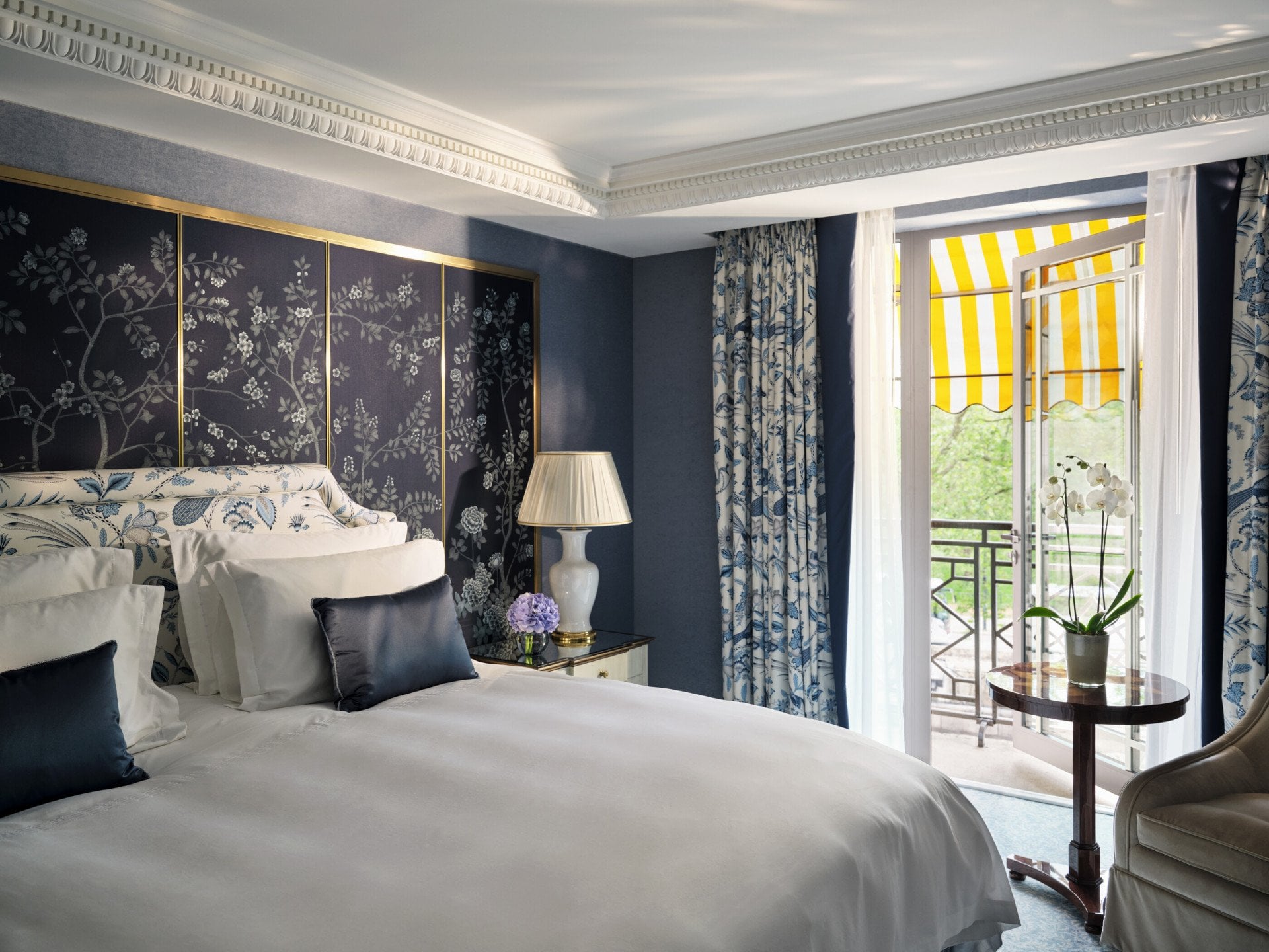 Bedroom in The Hyde Park Suite at The Dorchester, featuring blue floral décor, a king-sized bed and a private balcony with a yellow-striped awning.