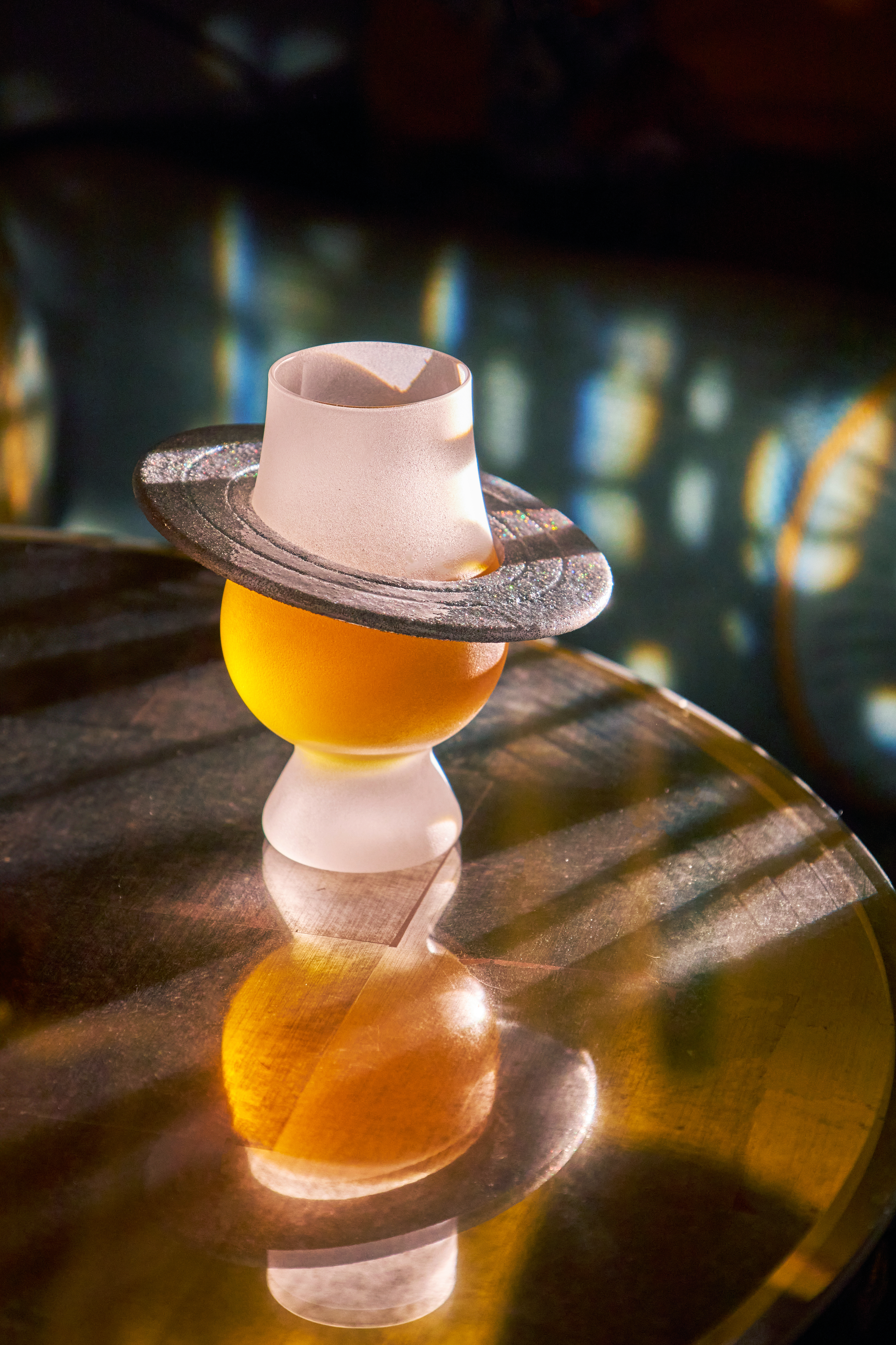 A cocktail from The Vesper Bar’s Planetarium menu, presented in a sculptural glass topped with a hat-shaped garnish on a reflective table.