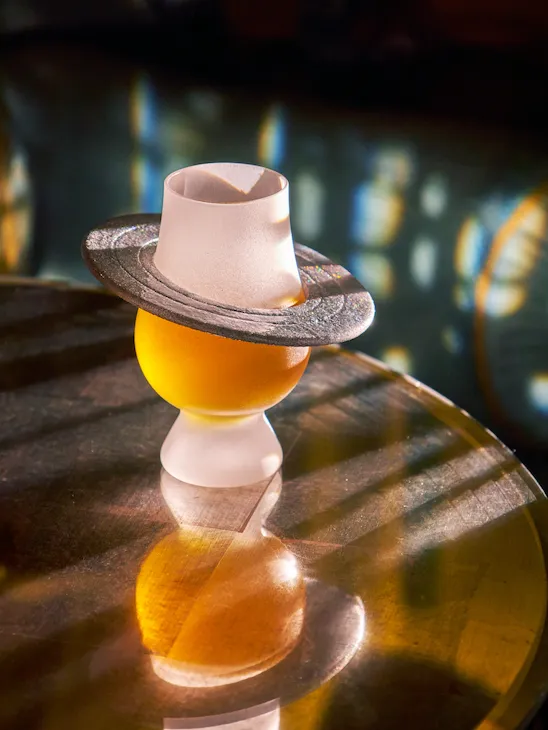 A cocktail from The Vesper Bar’s Planetarium menu, presented in a sculptural glass topped with a hat-shaped garnish on a reflective table.