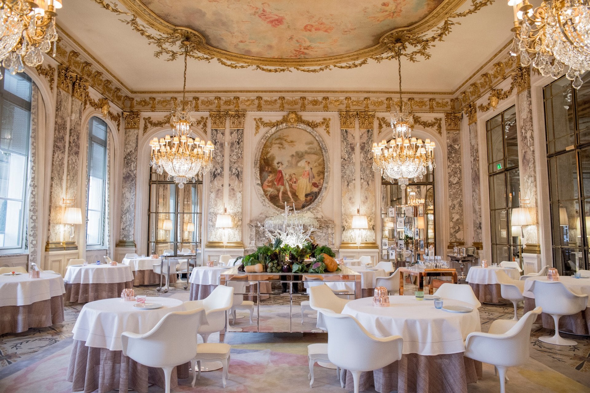 Luxurious dining room with ornate chandeliers, elegant round tables, and white chairs. Large murals and decorative gold accents create an opulent ambiance.