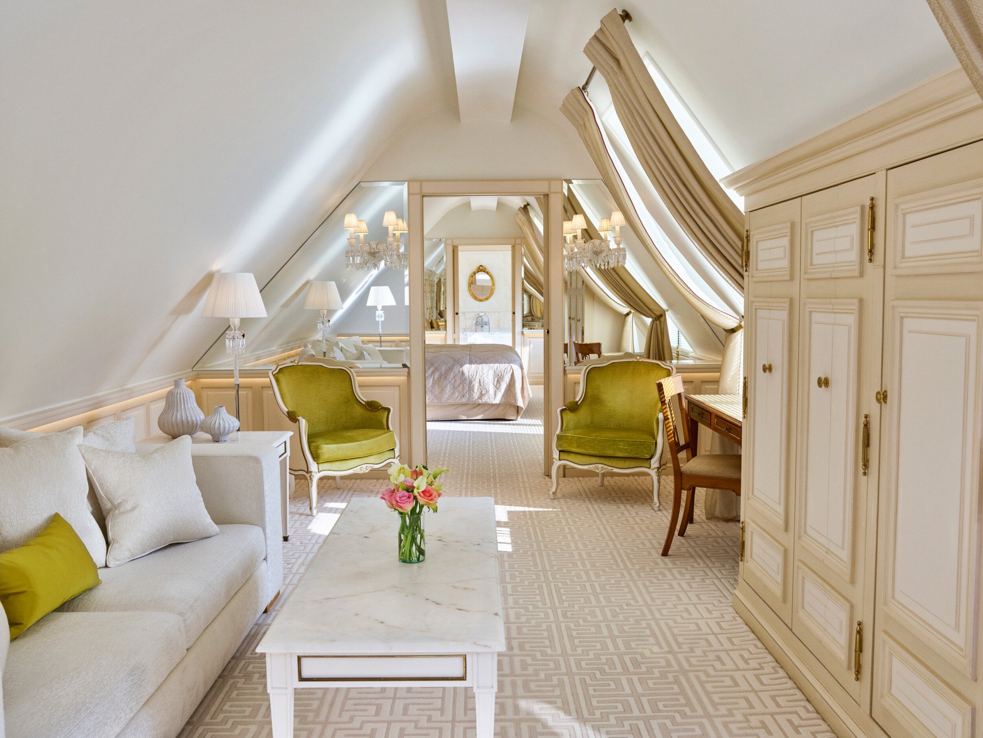 Chambre mansardée luxueuse avec plafond en pente, lustre élégant, décor crème et vert. Elle dispose d'un canapé confortable, d'une table en marbre et d'une lumière naturelle douce.