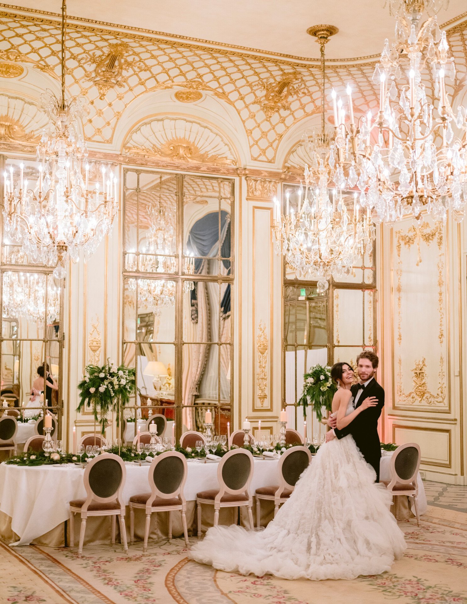 Wedding couple standing in front of a festive set table in Salon Pompadour. Green and white flower arrangements are on the table. The bride is laughing and in the arms of her husband, at Le Meurice, Paris