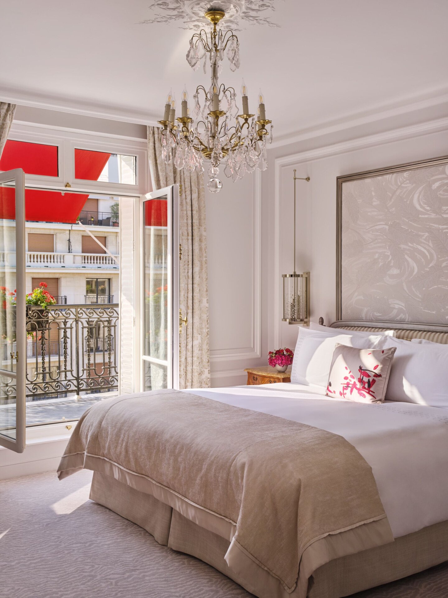 Quarto elegante com cama bege, lustre ornamentado e portas da varanda abertas revelando um toldo vermelho. Ambiente luminoso e arejado com decoração clássica.