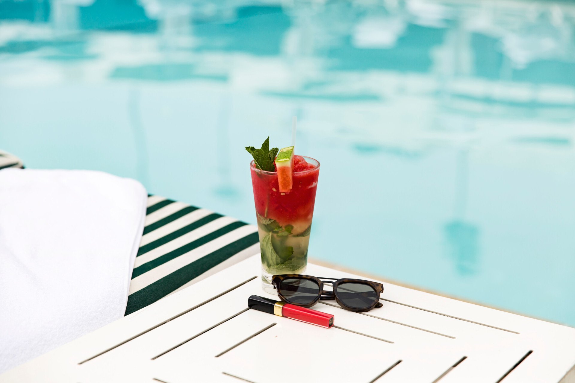 Colorful cocktail paired with sunglasses and lipstick on a table in front of the pool