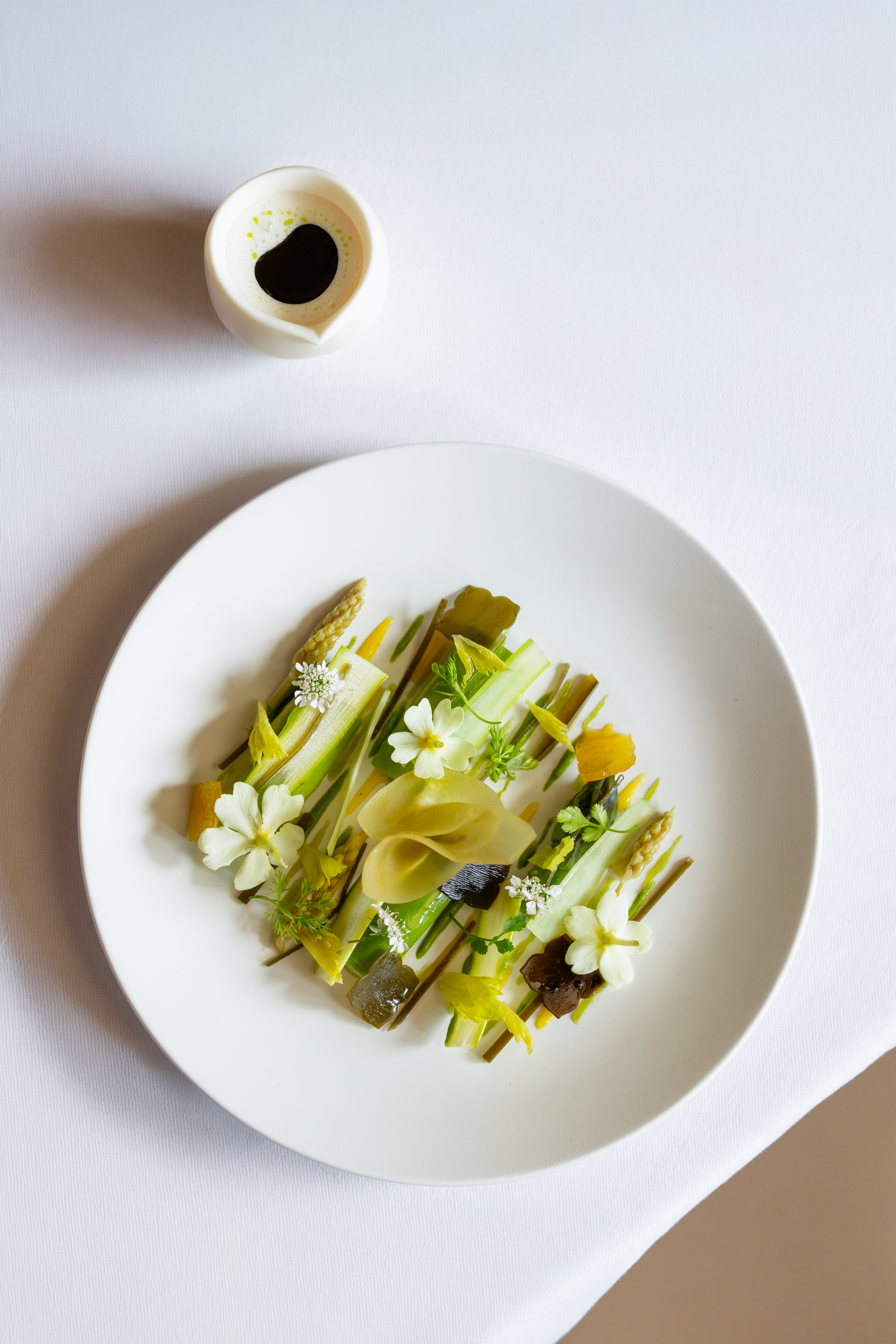 Elegant vegan pasta with herbs and flowers on a white plate, served with a small dish of soya sauce