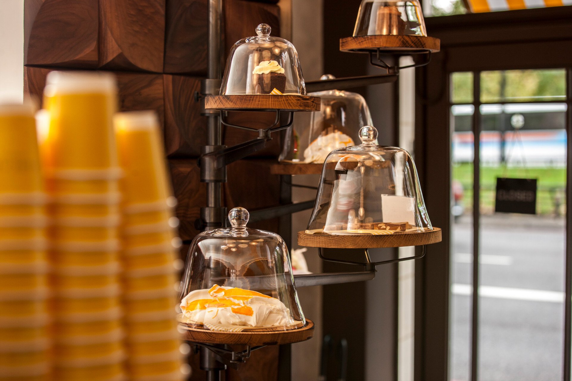Display of freshly prepared pastries at Parcafé at The Dorchester, showcasing sweet treats presented under glass cloches on tiered shelves, with takeaway coffee cups and views towards Hyde Park visible through the café window.