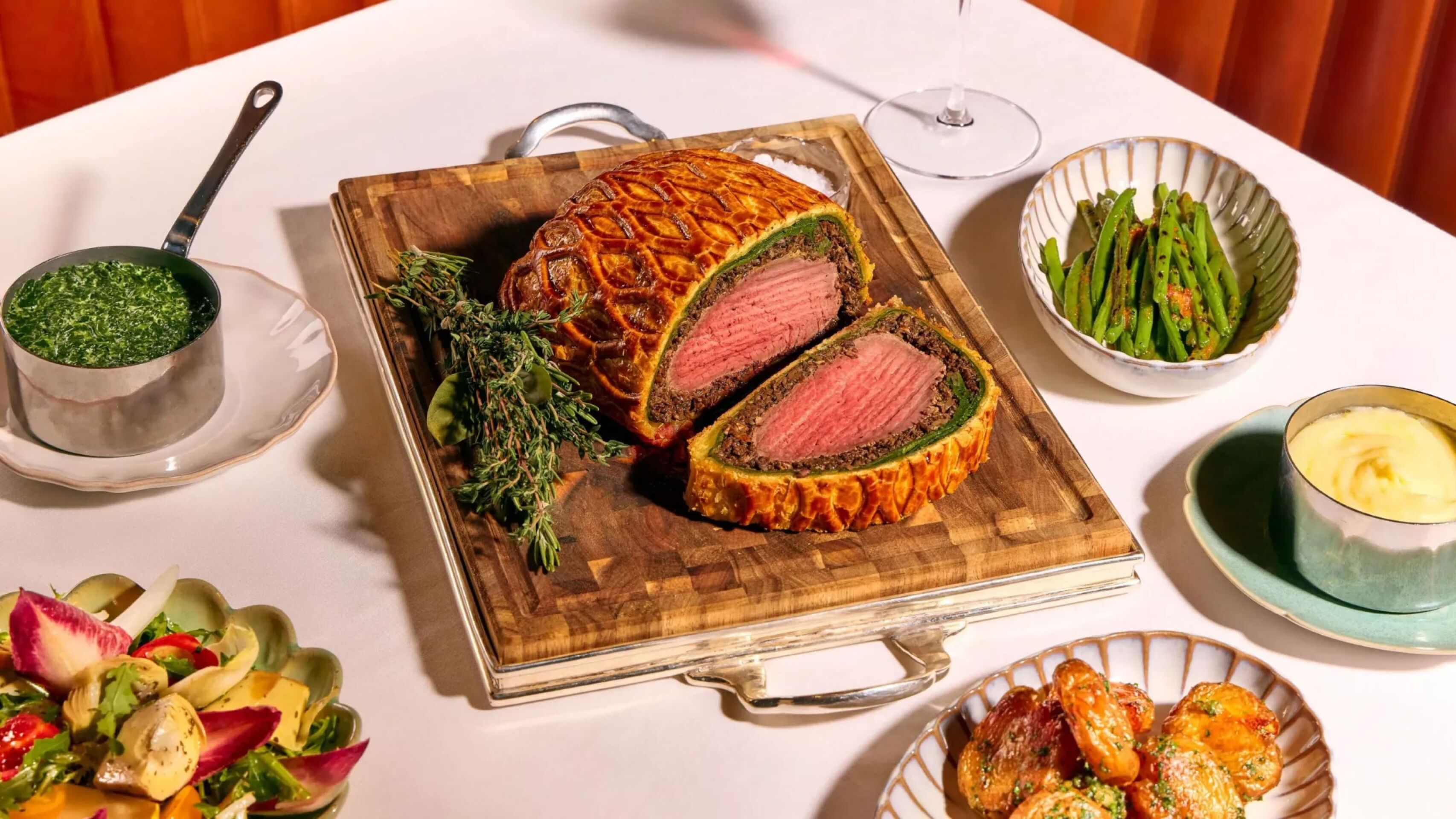 Dining table at The Grill at The Dorchester, showcasing a Beef Wellington sliced to reveal a pink centre, served with asparagus, creamed spinach, roasted potatoes and a fresh seasonal salad.