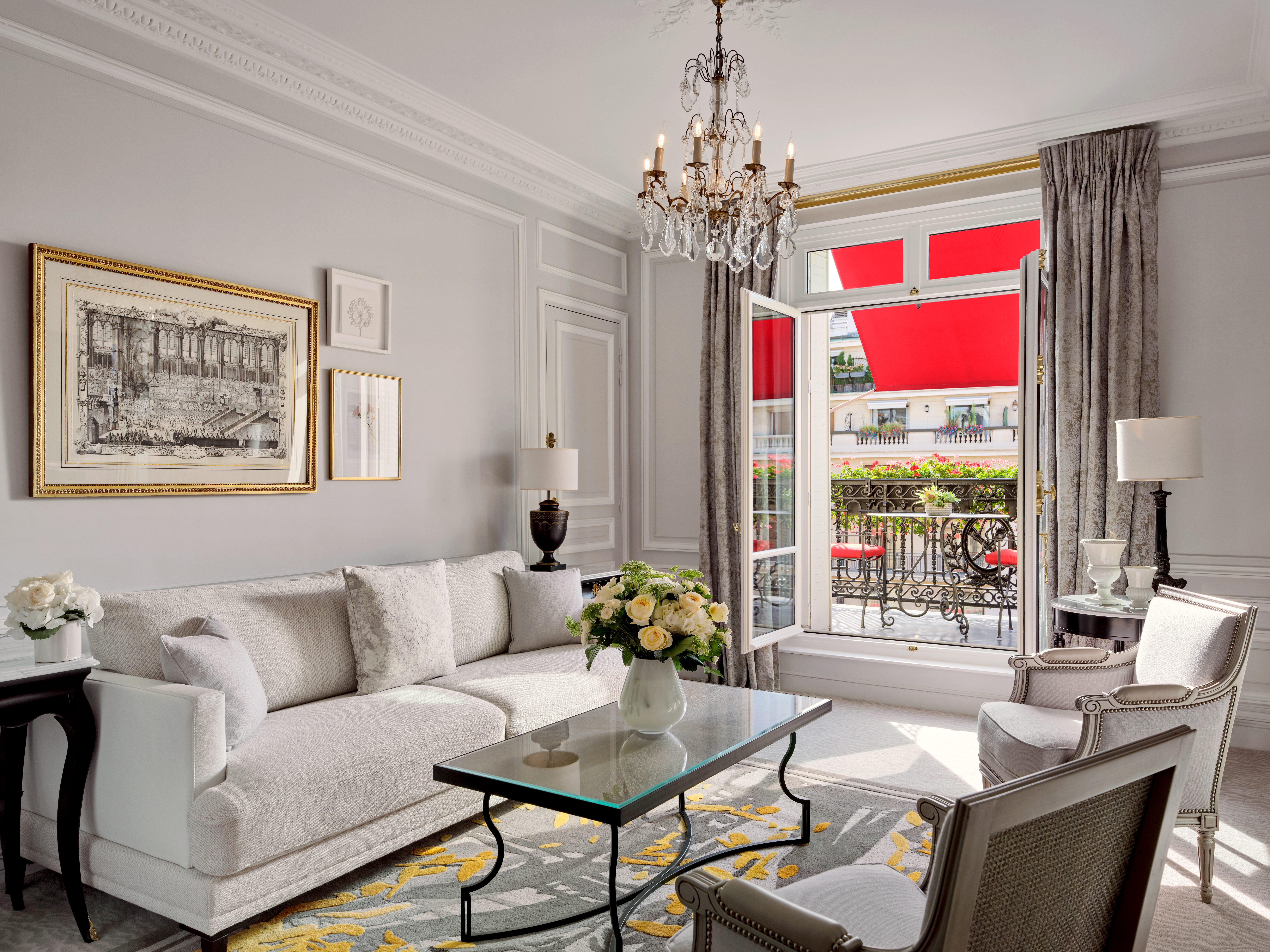 Salon élégant avec canapé blanc, table basse en verre et fauteuils. Un lustre est suspendu au plafond. Des portes ouvertes mènent à un balcon avec des auvents rouges.