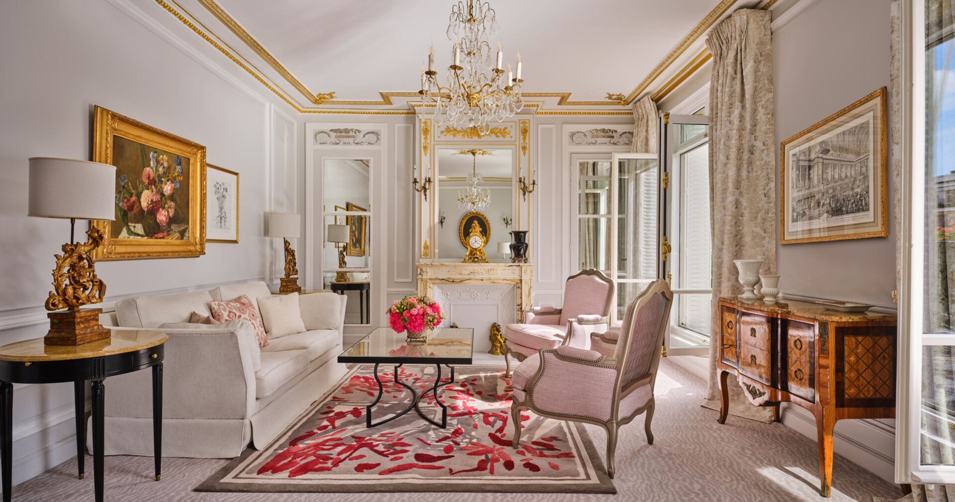 Salon élégant à la décoration classique : canapé blanc, fauteuils roses, lustre orné, touches dorées, compositions florales et tapis à motifs rouges. Ambiance chaleureuse et luxueuse.