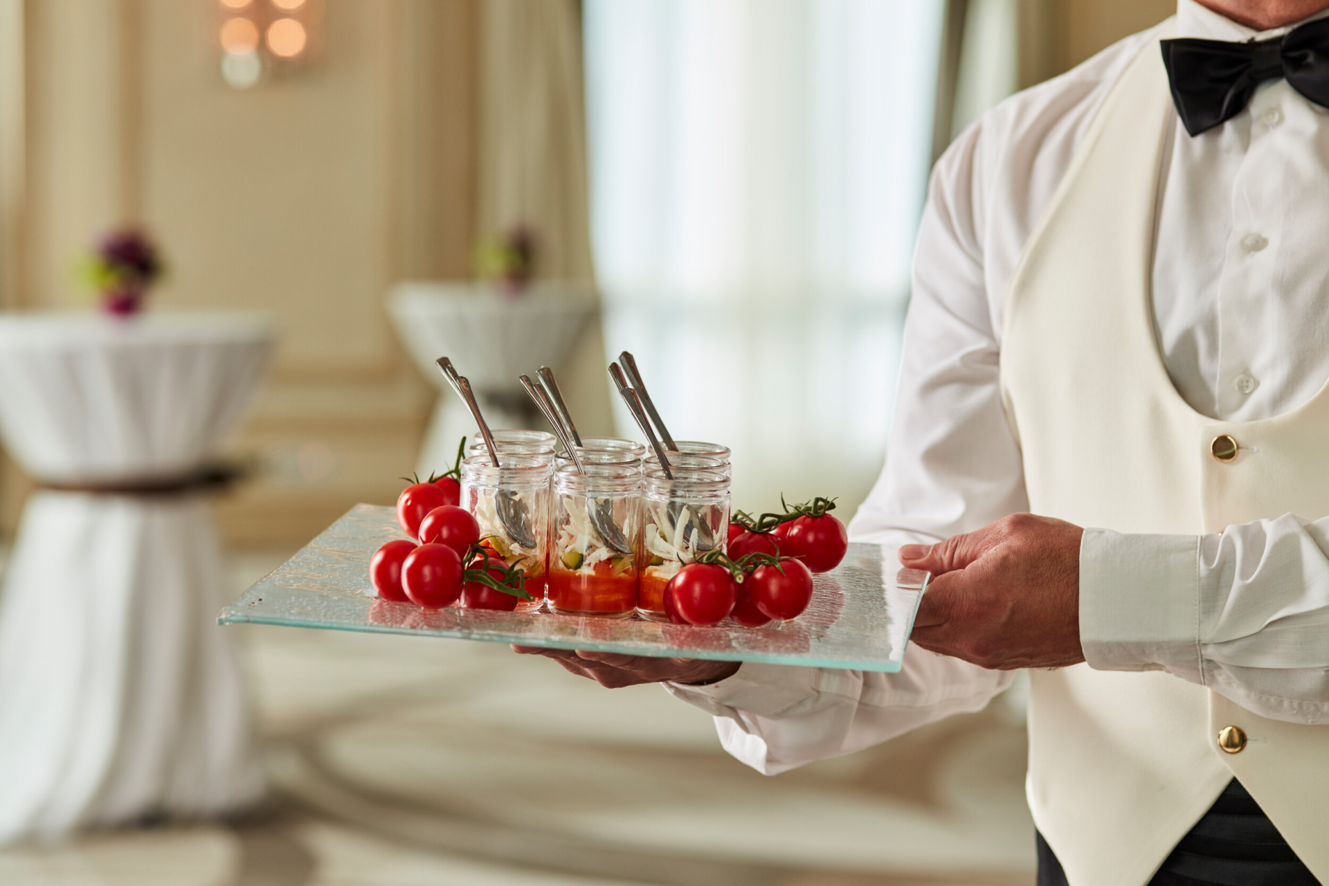 A close-up of a tray of canapés and decorative tomatoes being carried by a waiter. In the background, two high tables.