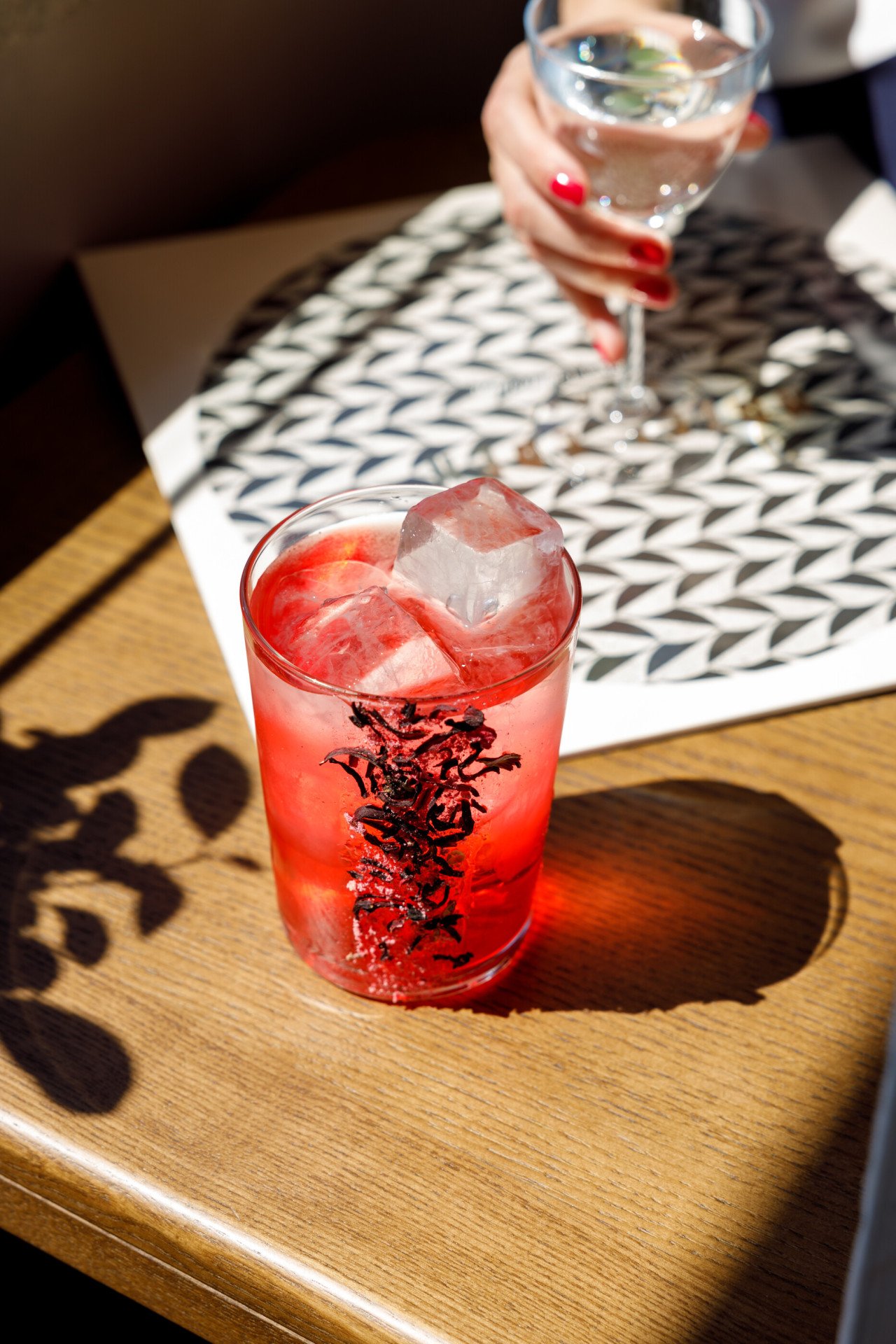 Cocktail rosso vivac guarnito con fiori di ibisco essiccati e grandi cubetti di ghiaccio. Il drink è posizionato su un tavolo di legno. Sullo sfondo, una donna tiene in mano un calice d'acqua sopra una tovaglietta con un motivo geometrico in bianco e nero.





