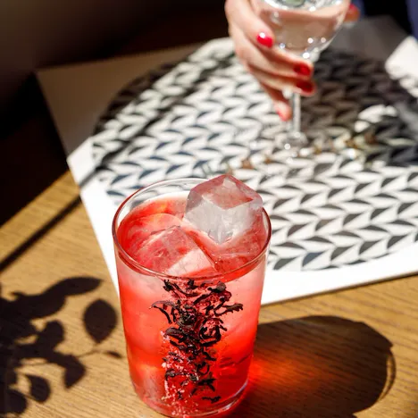Cocktail rosso vivac guarnito con fiori di ibisco essiccati e grandi cubetti di ghiaccio. Il drink è posizionato su un tavolo di legno. Sullo sfondo, una donna tiene in mano un calice d'acqua sopra una tovaglietta con un motivo geometrico in bianco e nero.