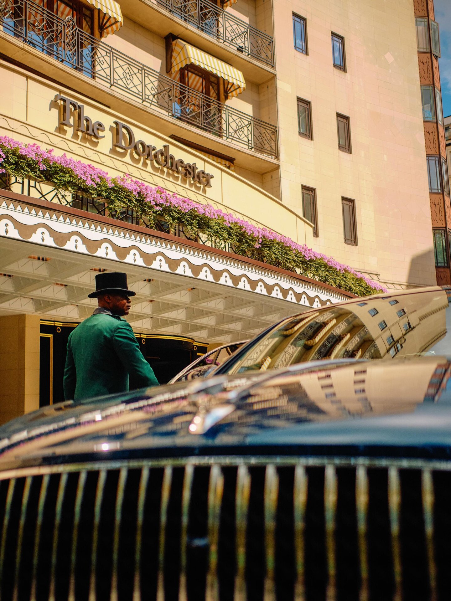 The Dorchester Bentley parked outside the hotel's main entrance on Park Lane, showcasing its distinctive grille and polished finish against the backdrop of The Dorchester's elegant façade, floral displays and signature signage.
