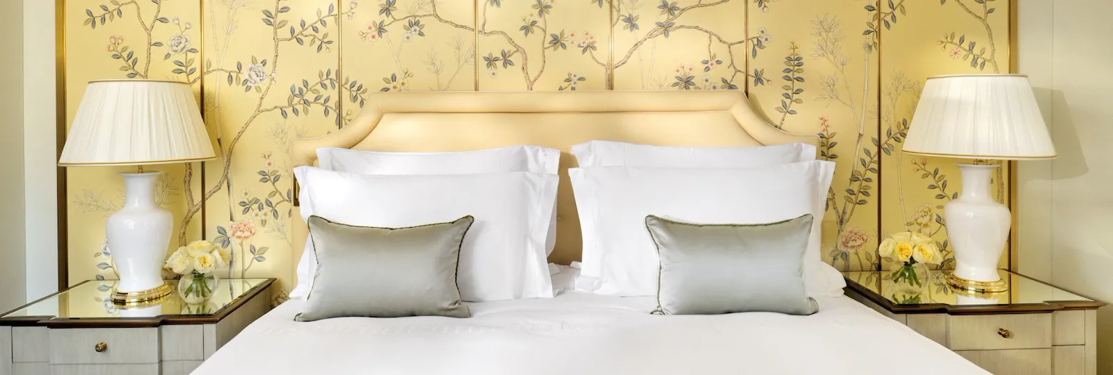 The Mayfair Suite bedroom at The Dorchester, showcasing a king-sized bed, elegant bedside tables and a decorative hand-painted wall panel featuring delicate flowering branches in soft gold and cream hues.