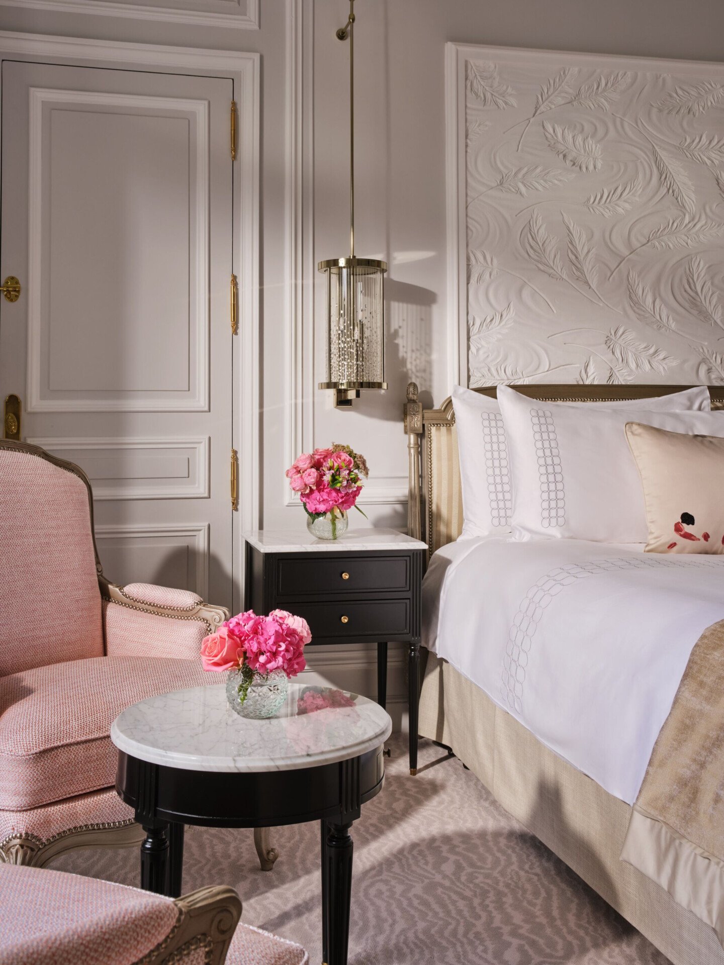 Um quarto luxuoso com uma poltrona rosa macia, uma pequena mesa com tampo de mármore e flores rosa, uma cama com lençóis brancos impecáveis e molduras elegantes nas paredes.