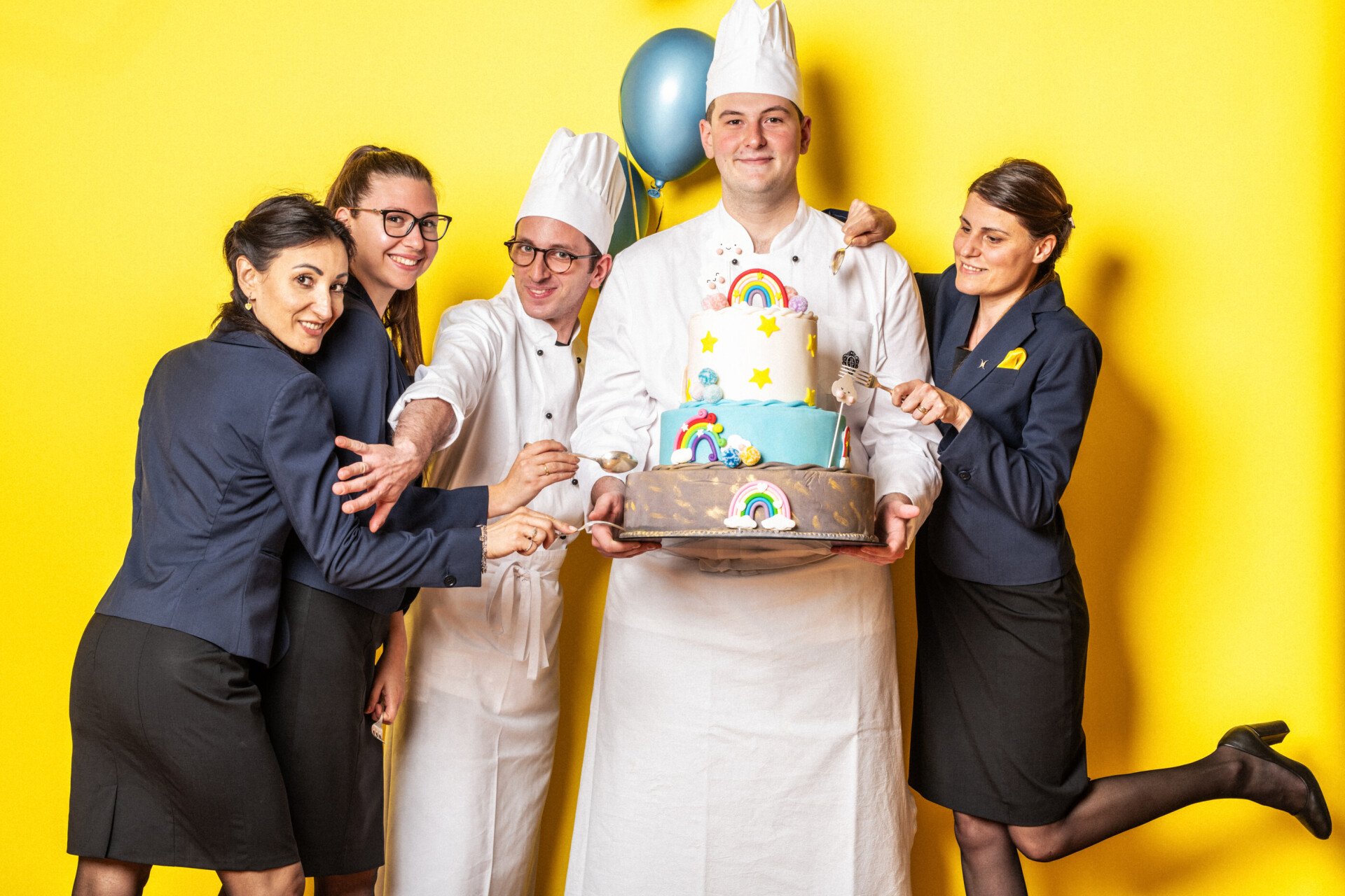 Photo of five employees against a yellow background, one holding a cake with rainbows