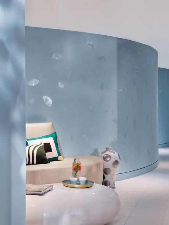 Salon moderne avec des murs bleu clair, un canapé beige orné de coussins géométriques et un grand vase en céramique. Une table basse brillante sur laquelle sont posées des boissons se trouve au premier plan.