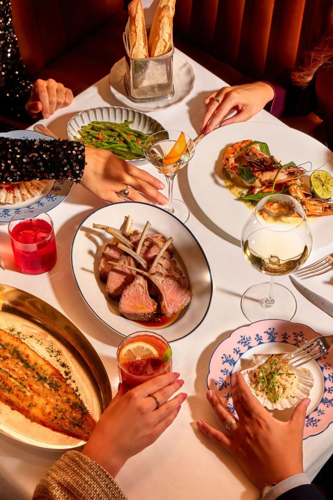 Dining table at The Grill at The Dorchester, showcasing a selection of signature dishes including sliced roast lamb, grilled prawns, dover sole, green beans, artisan bread and cocktails, presented for sharing.