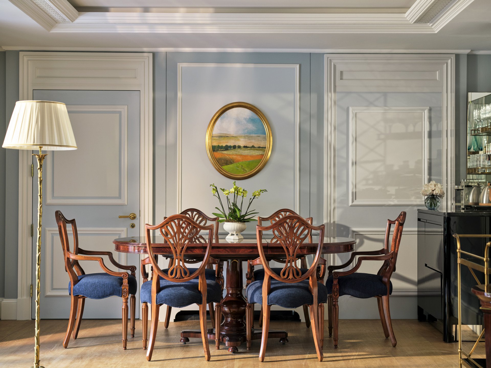 The Hyde Park Suite at The Dorchester, showcasing a formal dining area with a polished wooden table, blue-upholstered dining chairs, decorative wall panelling and an oval-framed landscape painting.