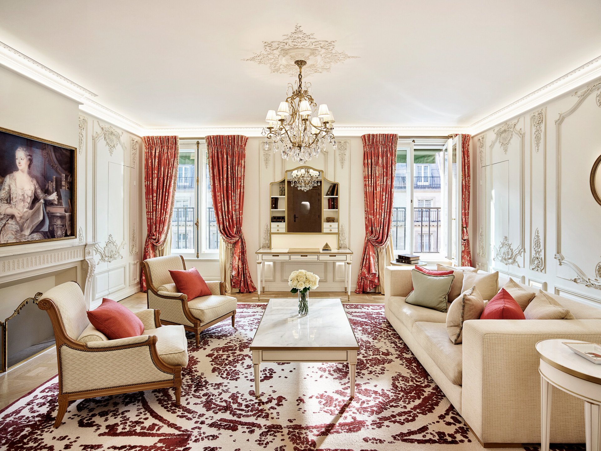 Salon luxueux avec lustre orné, canapés élégants et touches de rouge. Grandes fenêtres avec rideaux rouges, tapis à motifs floraux et tableau classique.