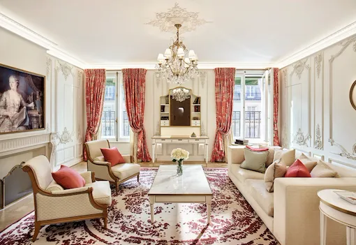 Sala de estar luxuosa com lustre ornamentado, sofás elegantes e detalhes em vermelho. Janelas amplas com cortinas vermelhas, tapete floral e pintura clássica.