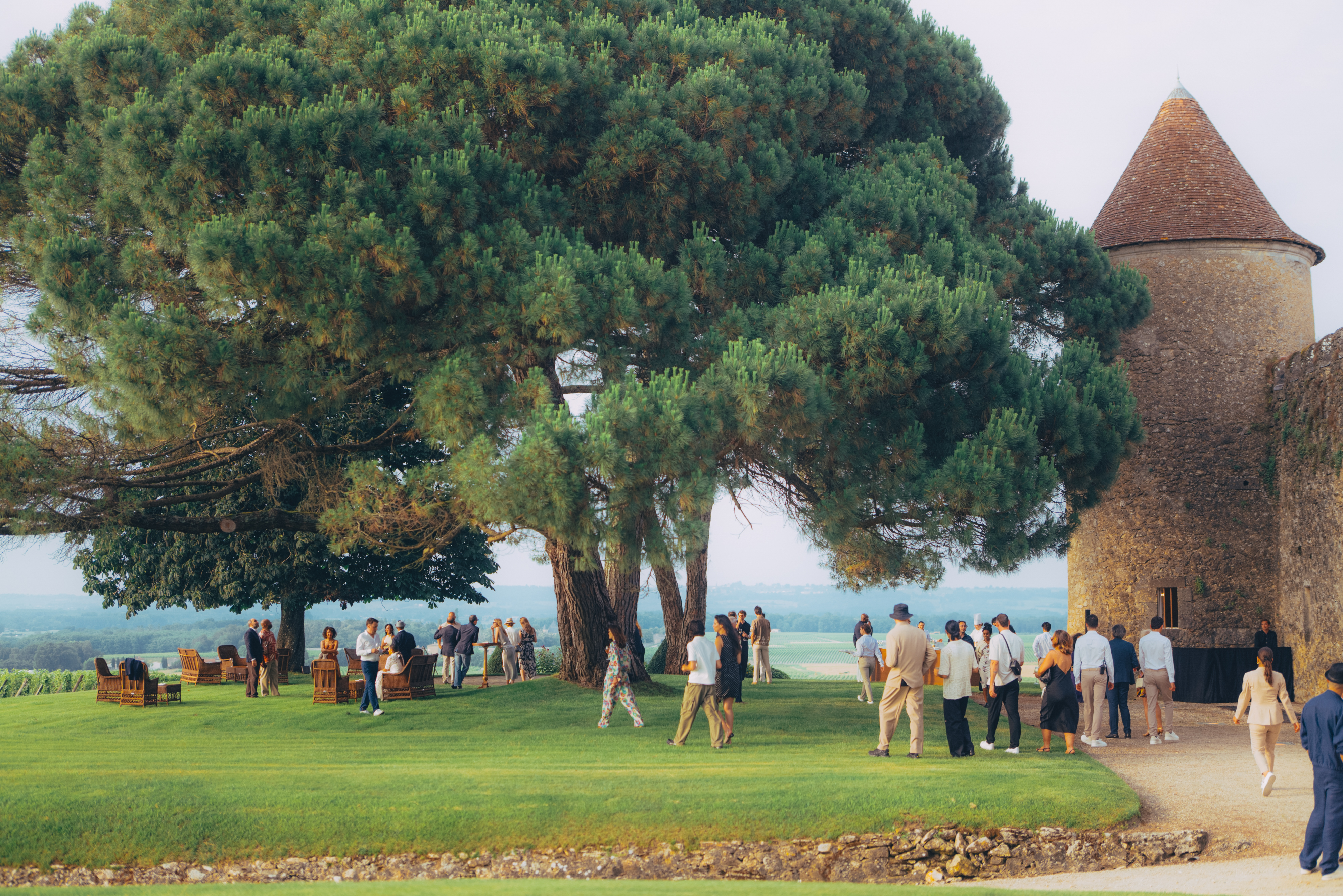 A group of people gather on a lush green lawn under large, shady trees next to an old stone tower. The scene is relaxed and social, with clear skies.