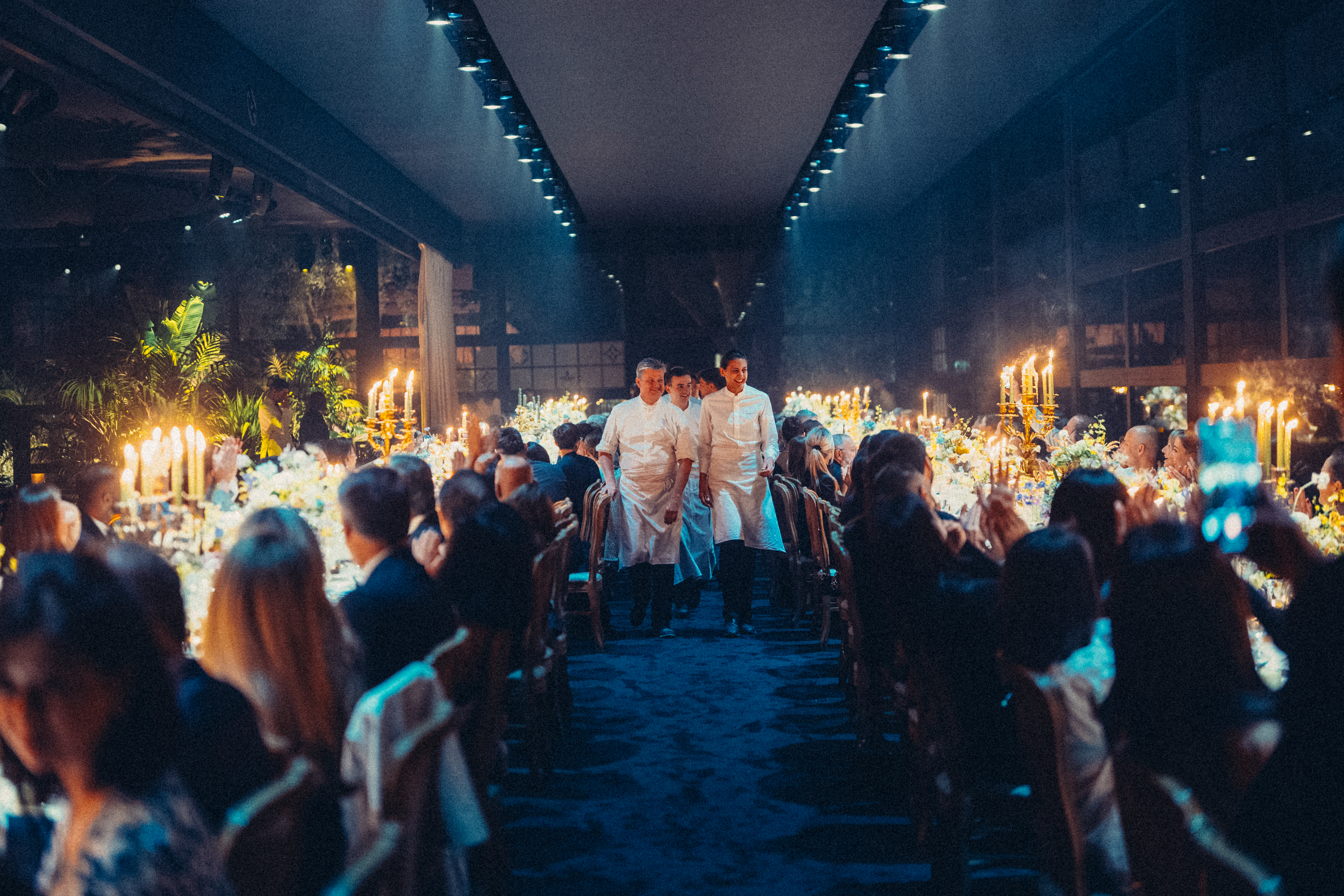 Elegant banquet scene with chefs walking between long tables set with candles and flowers. Guests are seated, creating a warm, festive atmosphere.