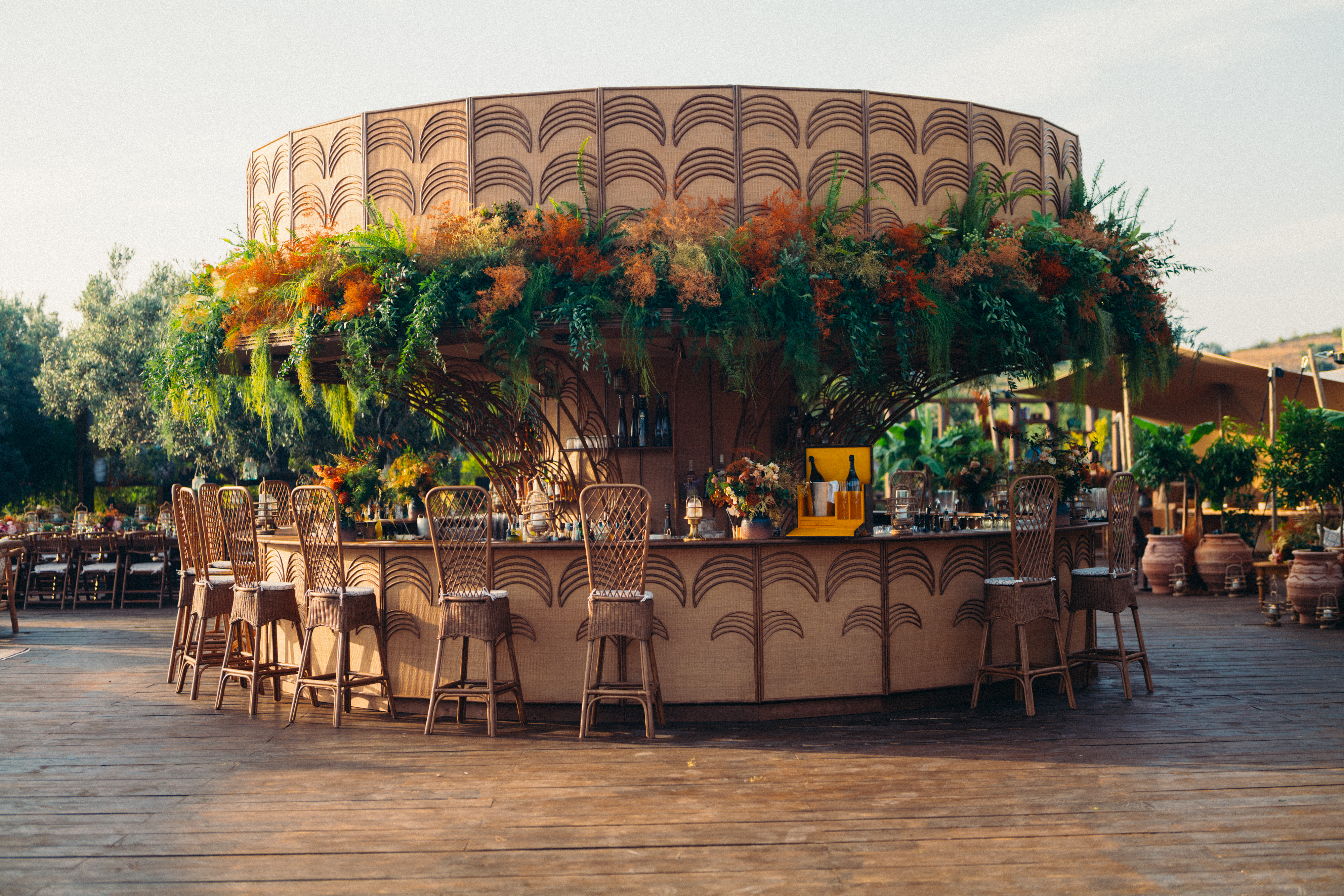 A round outdoor bar features tall wicker chairs and lush green and orange foliage. It stands on a wooden deck amid an inviting, sunlit garden setting.