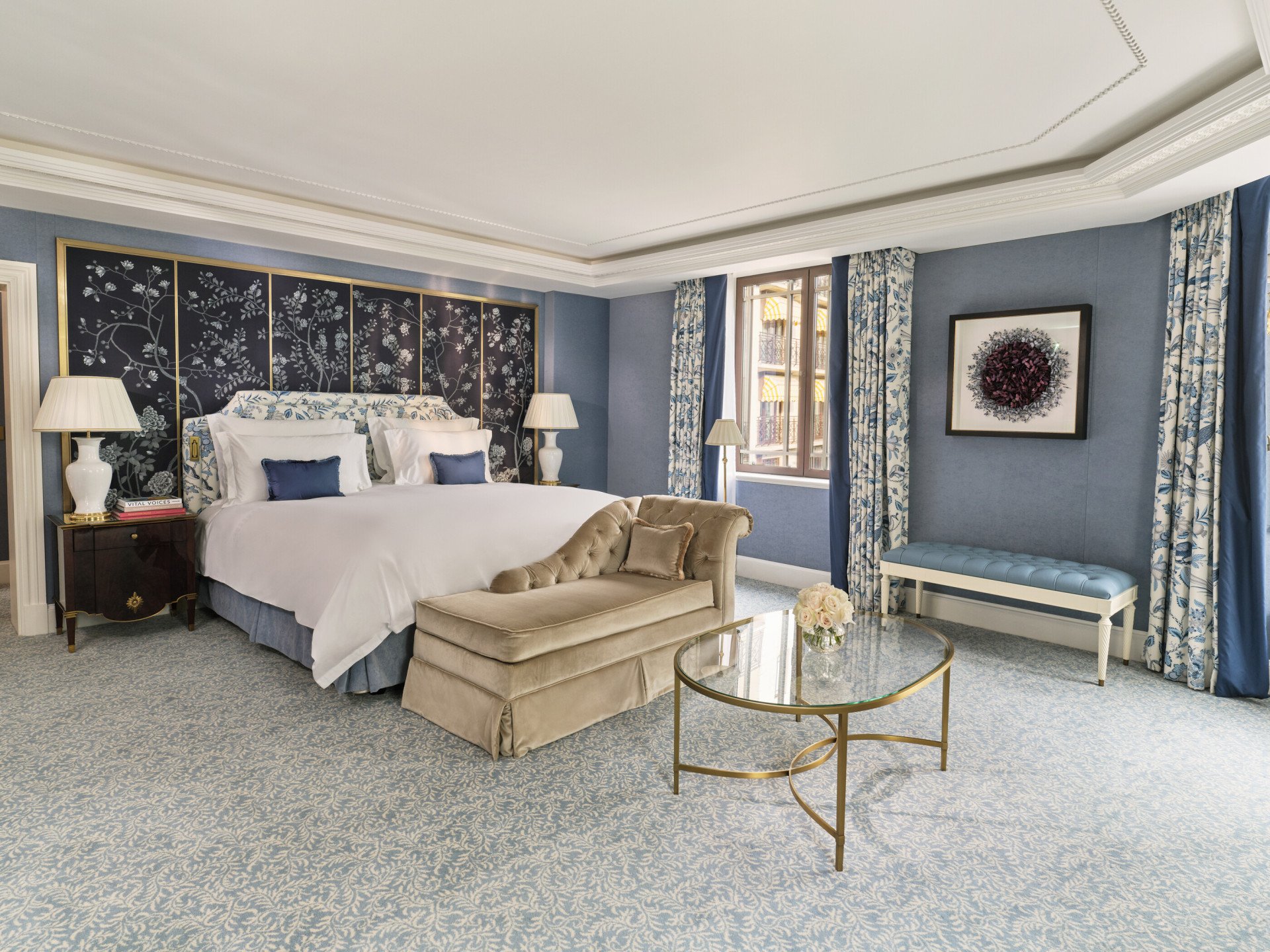 The Regal Suite bedroom at The Dorchester, showcasing soft blue walls, an upholstered king-sized bed, decorative chinoiserie-style wall panels, a seating area and large windows filling the room with natural light.