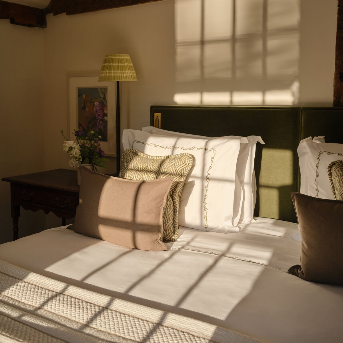 Sunlit bedroom with exposed beams and green velvet headboard in a cottage room at Coworth Park, Ascot