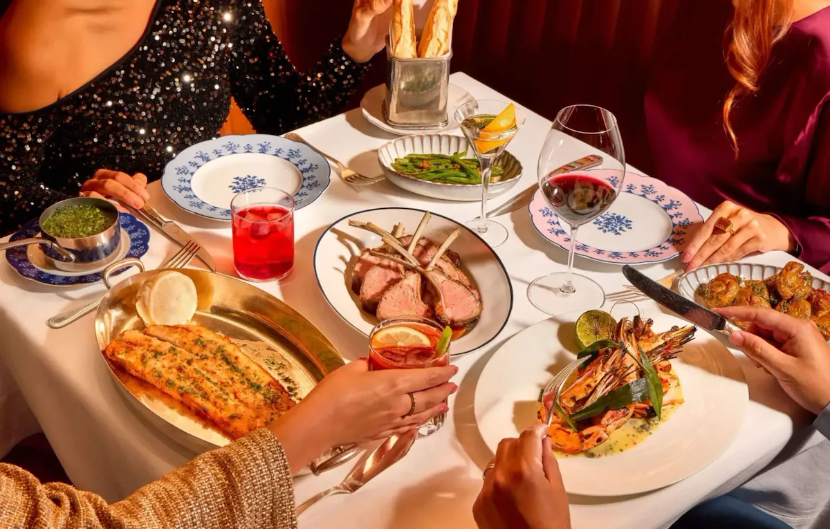 Overhead view of a dining table at The Grill at The Dorchester with lamb chops, grilled prawns, Dover sole, green beans, sauces, cocktails and guests serving themselves from shared dishes.