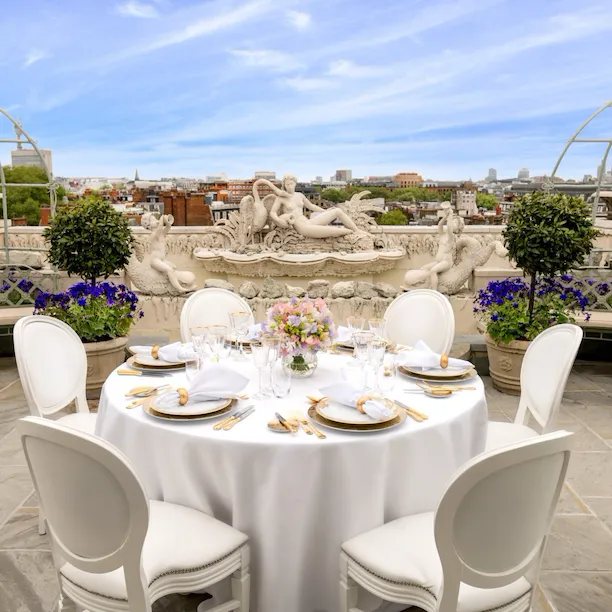 Luxurious rooftop terrace at The Penthouse & Pavilion at The Dorchester, showcasing a round table elegantly laid for dining, surrounded by white chairs, seasonal flowers, classical stone statuary, manicured topiary and panoramic views of the London skyline beneath a blue sky.