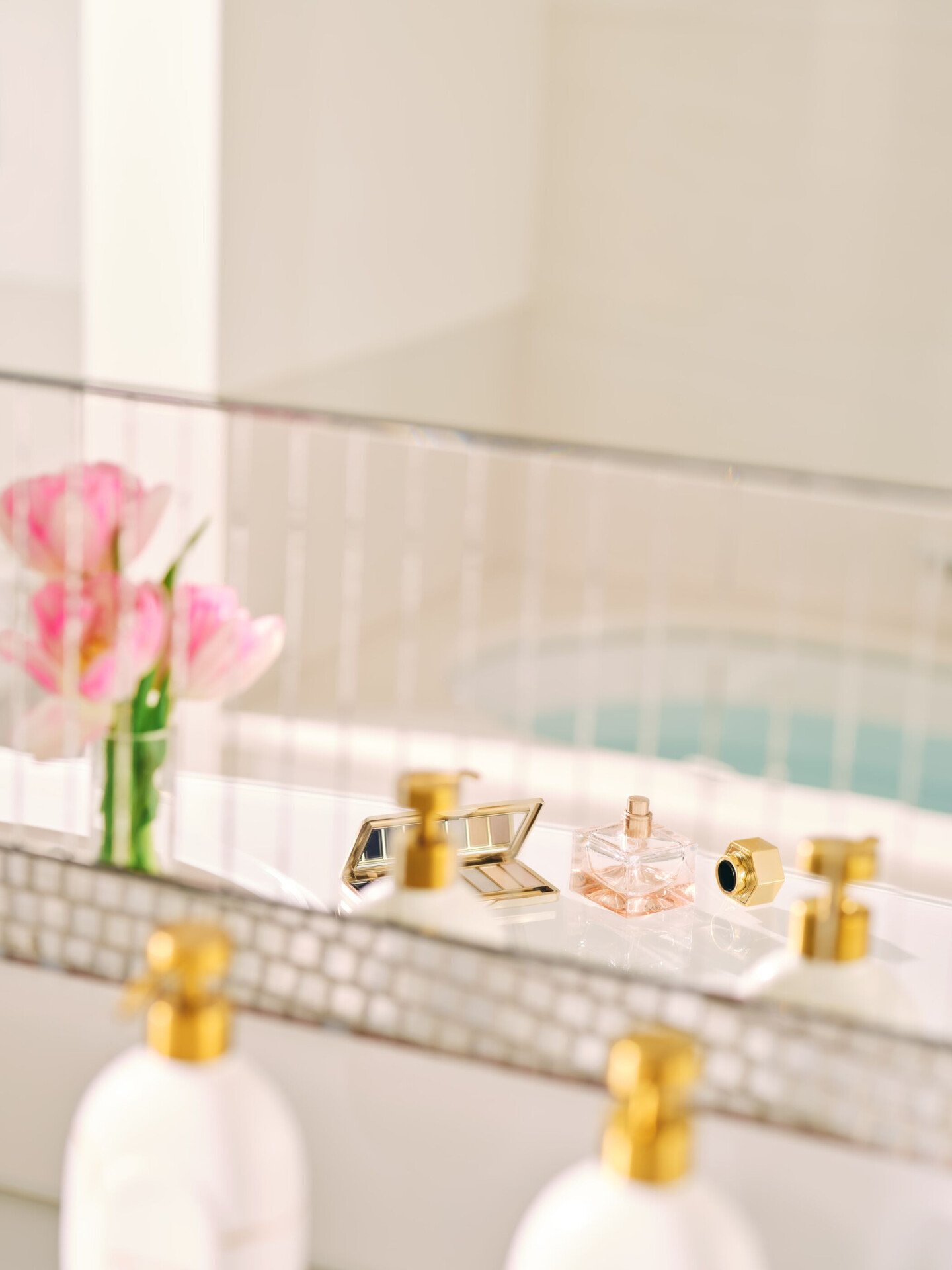 riflesso dello specchio di un bagno con fiori rosa, un profumo, una palette di trucchi e dei dispenser di shampoo