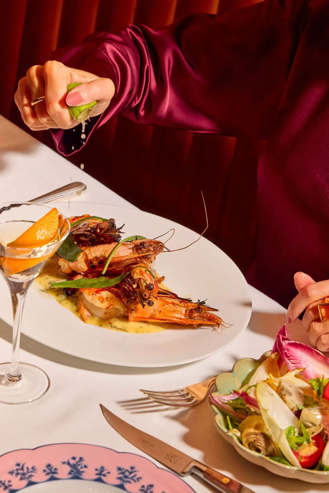 Guest squeezing fresh lime over grilled prawns at The Grill at The Dorchester, served with a Martini cocktail and seasonal salad.