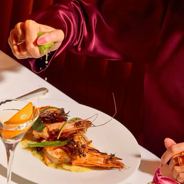 Guest squeezing fresh lime over grilled prawns at The Grill at The Dorchester, served with a Martini cocktail and seasonal salad.