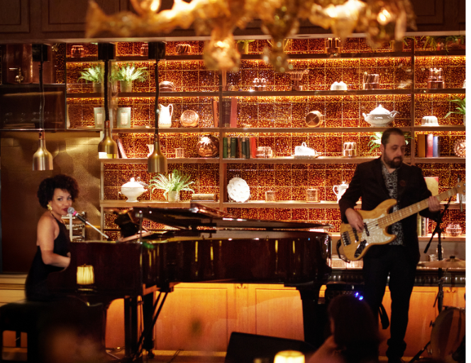 Ronnie Scott’s at The Dorchester, showcasing a live performance with musicians playing piano and bass, framed by warm lighting and backlit shelves displaying decorative objects and glassware.