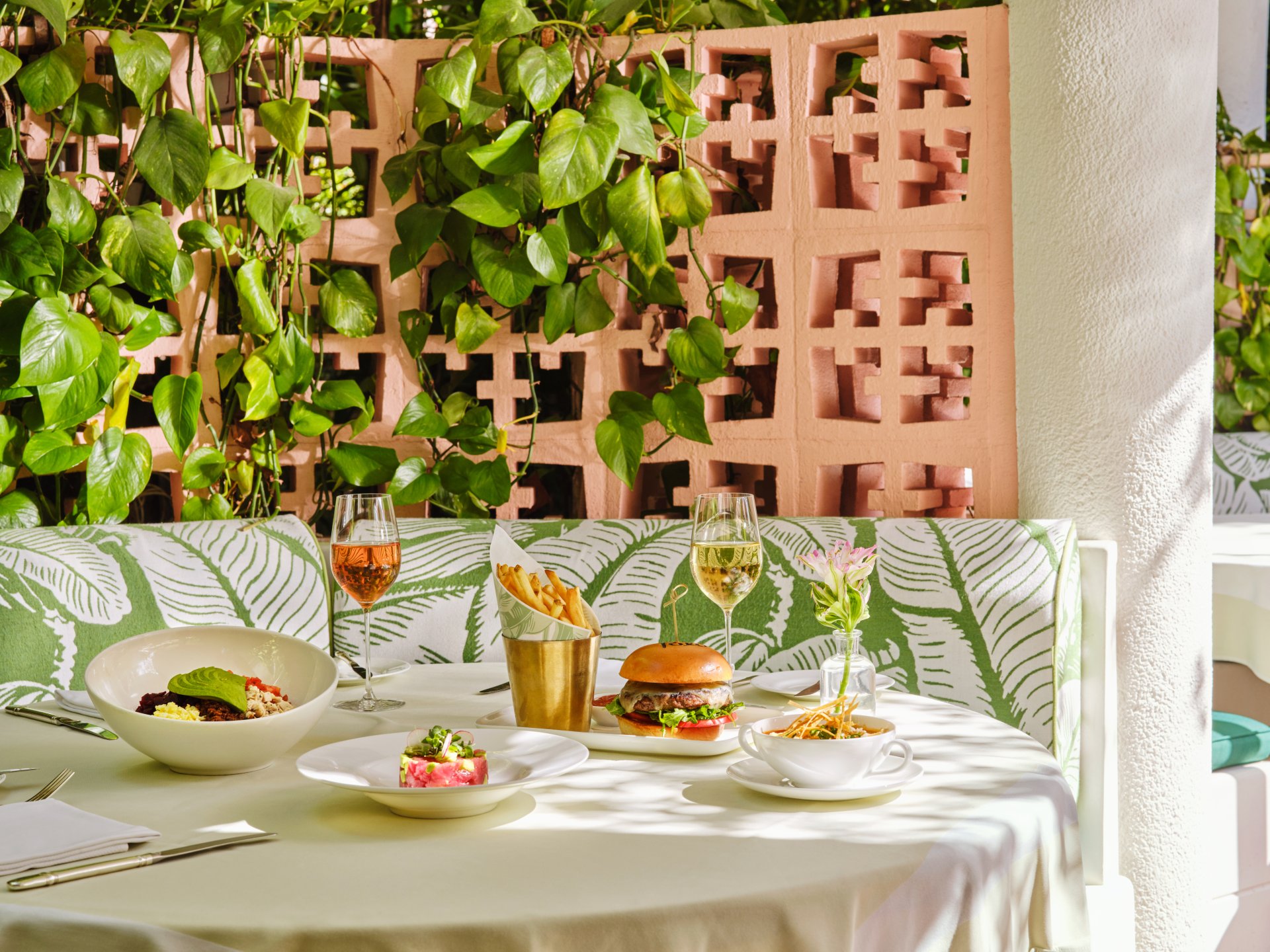 Outdoor table set with drinks, food, and leafy decor in bright, relaxed atmosphere