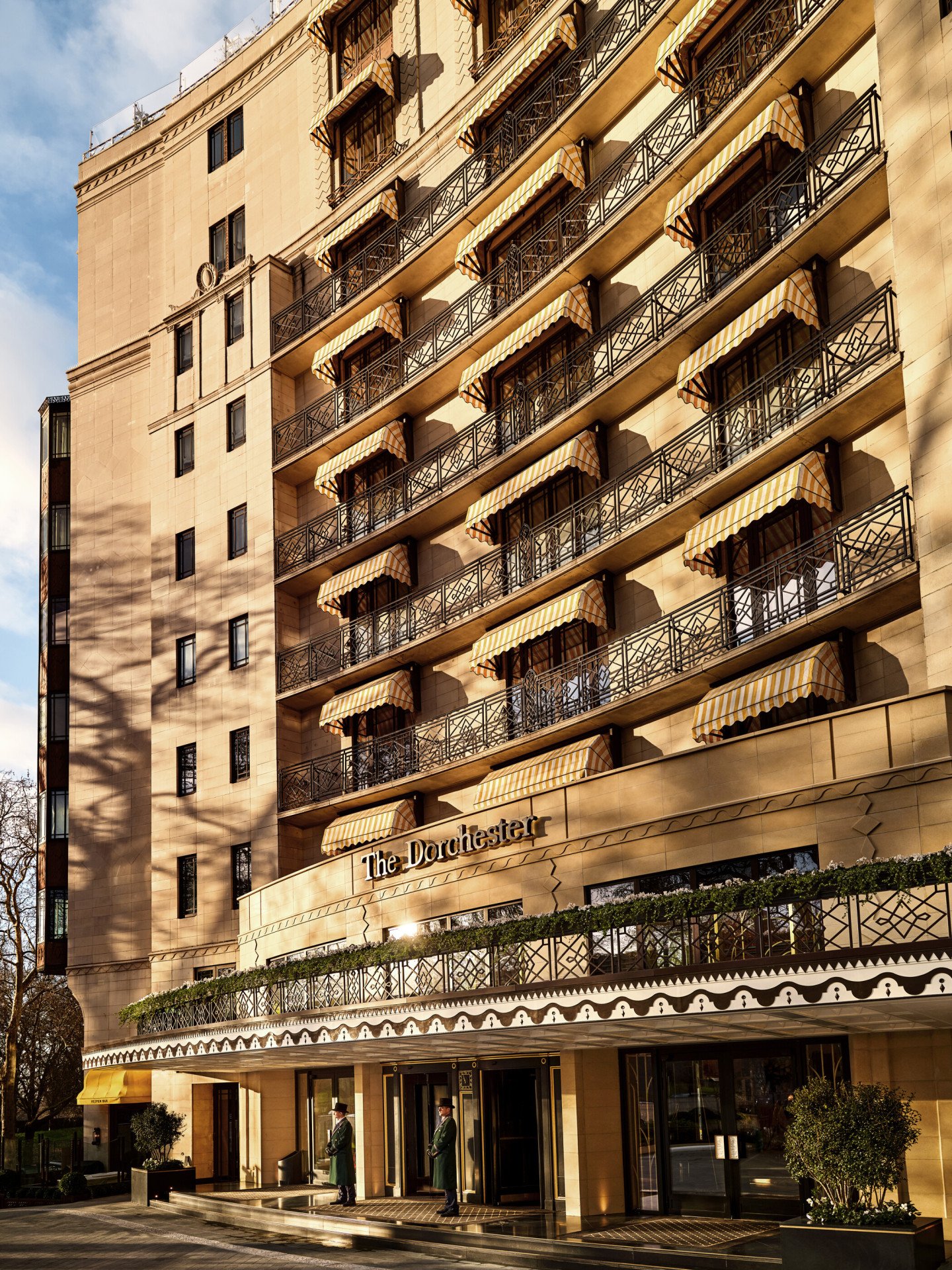 Exterior view of The Dorchester, showcasing its elegant Park Lane façade, decorative wrought-iron balconies, signature yellow-striped awnings and the hotel's welcoming entrance bathed in warm sunlight.