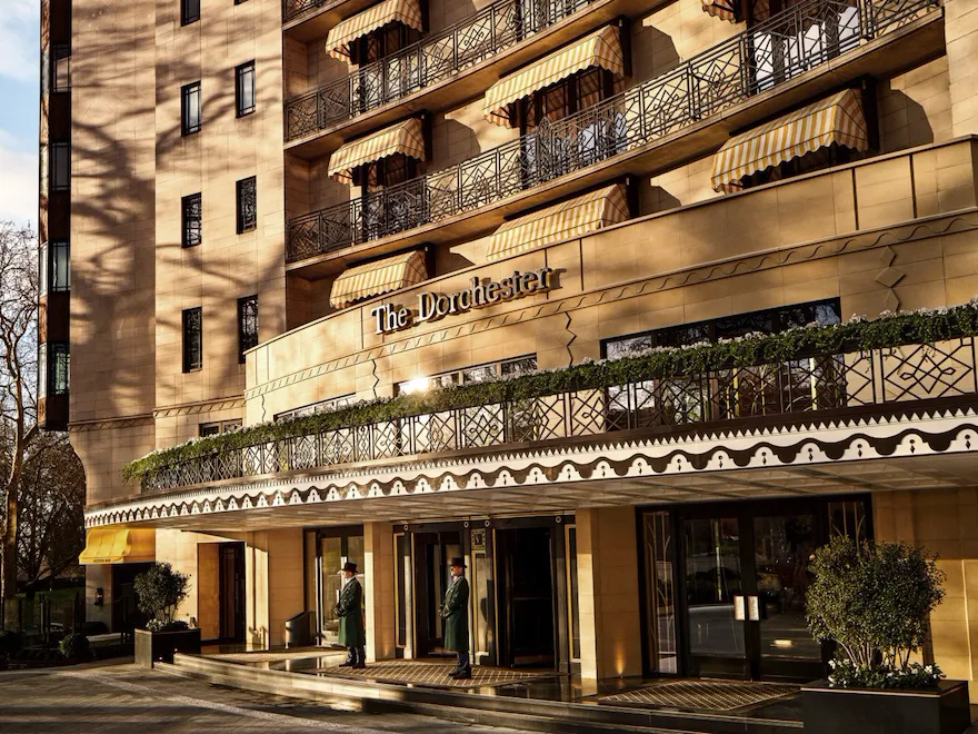 Exterior view of The Dorchester, showcasing its elegant Park Lane façade, decorative wrought-iron balconies, signature yellow-striped awnings and the hotel's welcoming entrance bathed in warm sunlight.